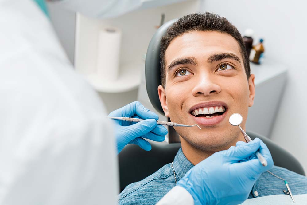 A dental professional is preparing a patient s teeth with instruments while the patient smiles.