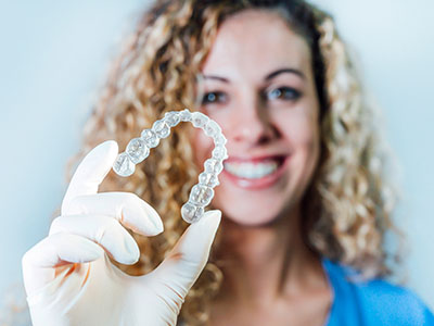 A woman wearing a blue surgical mask holds up a transparent plastic tray containing a clear dental implant.