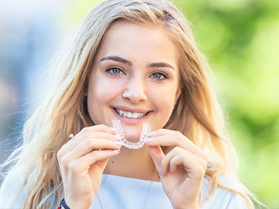 A young woman holding up a clear plastic retainer with a smile, showcasing her teeth alignment.