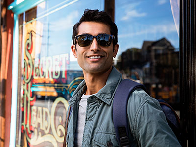The image features a man standing outdoors in front of a storefront window with a sign that reads  Sunday s  on it, wearing sunglasses, a backpack, and a jacket, posing for the camera with a smile.