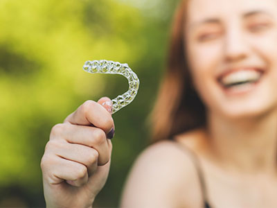 The image features a smiling individual holding up a clear plastic dental impression mold with a toothy grin, against a blurred outdoor background.