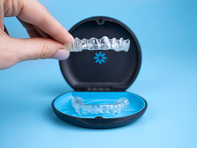 A hand holding a clear plastic dental retainer with visible teeth impressions, placed inside an open blue case with a white circular logo on the lid.