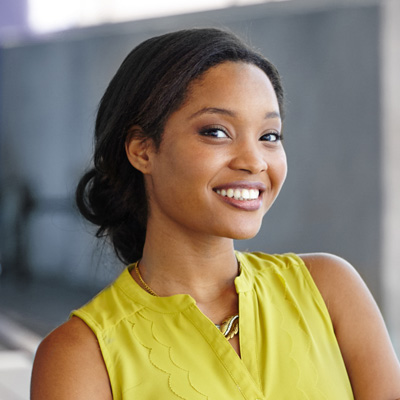 The image features a smiling woman with dark hair, wearing a yellow top, posing against a blurred background that suggests an urban setting.