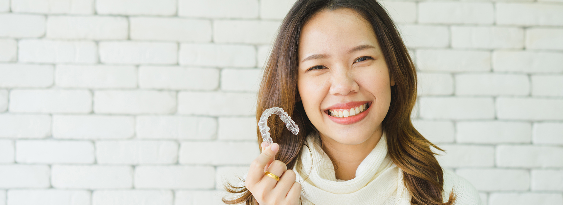 A woman with a radiant smile holds up a ring, standing against a brick wall.