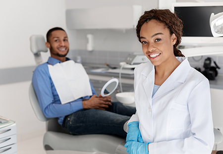 A dental professional in a white coat smiling at a patient seated in a dental chair, with a man sitting behind them observing.